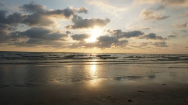 Altın gün batımı nemli kumda büyüleyici bir yansıma yaratır. Yumuşak dalgalar kıyıya doğru yuvarlanırken bulutlu bir gökyüzünün altında sahil şeridinin huzurlu ve pitoresk bir görüntüsünü sunar.