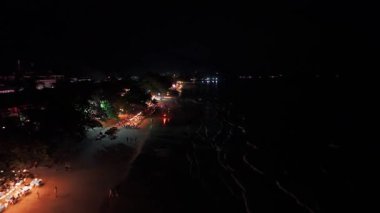 Yumuşak dalgalar, turistler deniz kenarındaki restoranların parıldayan ışıkları altında yemek yerken kumlu sahili nazikçe okşayarak canlı ve büyülü bir atmosfer yaratıyor.