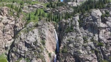 Berrak su zarif bir şekilde sarp, kayalık bir dağ yamacından aşağı akar, yemyeşil bitki örtüsünün ortasında güzel bir manzara sunan pitoresk bir şelale yaratır.