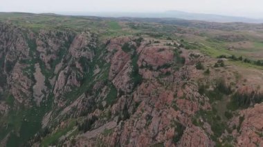 Görkemli dağ arazisi engebeli kayalık araziyi gözler önüne seriyor, yoğun olarak canlı yeşil bitki örtüsüyle kaplı, dramatik bulutlu ufuk çizgisinin altında geniş bir vahşi doğa manzarası var.