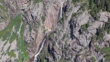 Kristal berrak su dik kayalık dağ yamacından aşağı akıyor, çarpıcı jeolojik oluşumuyla el değmemiş el değmemiş vahşi doğada canlı yeşil bitki örtüsüyle çevrili.