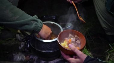 Avcı grubu tencereden güveci paylaşıyor, huzurlu vahşi gece boyunca parlayan kamp ateşinin yanındaki ahşap kaselere döküyor, kırsal alanda yemek sahnesini aydınlatıyor.