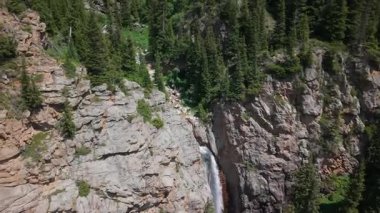 Berrak mavi gökyüzünün altında yemyeşil çam ormanlarıyla çevrili çağlayan bir şelalenin yer aldığı kayalık bir dağ manzarası nefes kesici.
