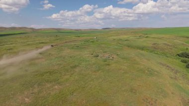 Siyah araba tozlu toprak bir yolda uçsuz bucaksız yeşil bir çayırda hızla ilerlerken arkasında beyaz bulutlarla benekli parlak mavi bir gökyüzünün altında uzun bir toz bulutu bırakıyor.