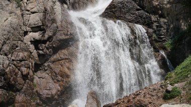 Artan saf beyaz su engebeli granit kayalıklardan aşağı akıyor, yoğun bitki örtüsüyle çevrili, saf güzellik ve gücü ön plana çıkarıyor.