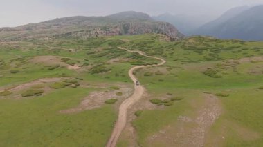 Koyu renkli off-road aracı yeşil dağ çayırlarında dolambaçlı toprak yoldan geçiyor engebeli manzarayı ve bulutlu yaz gökyüzünün altındaki el değmemiş yabanı ortaya çıkarıyor.
