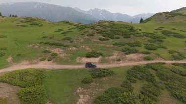 Yeşil dağ vadisinde dolambaçlı toprak yolda ilerleyen siyah SUV, uzak tepelere doğru uzanan el değmemiş el değmemiş yaban hayatıyla panoramik manzarayı gözler önüne seriyor.