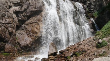 Artan saf beyaz su engebeli granit kayalıklardan aşağı akıyor, yoğun bitki örtüsüyle çevrili, saf güzellik ve gücü ön plana çıkarıyor.