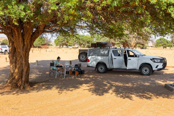 stock image Sesriem Campsite, Sossusvlei, Namibia - 01 October 2018: A girl sitting under an acacia tree at the campsite. Car with equipment parked nearby.