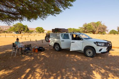 Sesriem Kampı, Sossusvlei, Namibya - 01 Ekim 2018: Kamp alanındaki bir akasya ağacının altında oturan bir kız. Yakınlarda park edilmiş bir araba..