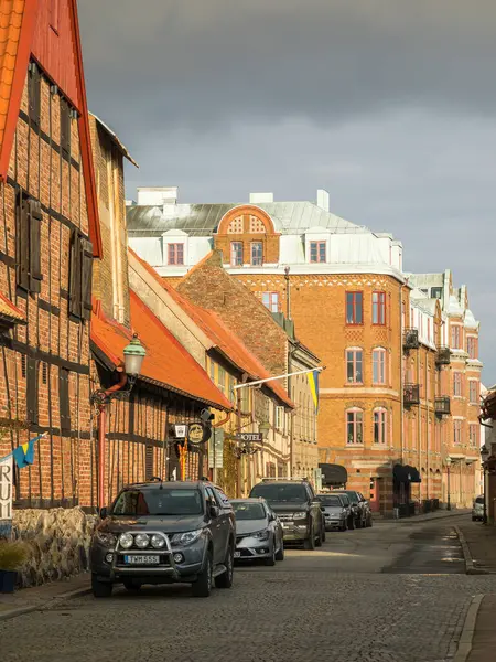 Ystad, İsveç - 7 Mart 2020: Bulutlu gökyüzü altında park edilmiş arabaları ve tarihi tuğla mimarisi olan bir kaldırım taşı caddesi.