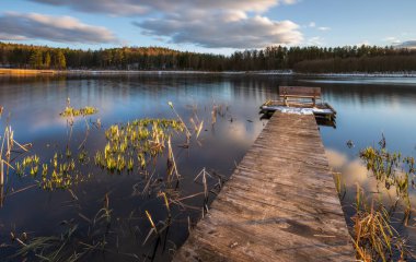 Polonya 'nın Okuniewo kentindeki ormanla çevrili durgun bir göl üzerinde ahşap iskele ve bank bulunan sakin bir bahar manzarası..