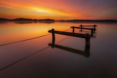 Ahşap iskeleleri ve yansımaları olan sakin bir gölün üzerinde renkli bir gün batımı, Rydzewo, Mazury, Polonya.