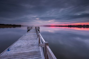 Polonya 'nın Mazury kentindeki sıcak ve renkli bir gökyüzü tarafından çerçevelenmiş, gün doğumunda sakin bir göle uzanan ahşap bir iskele..