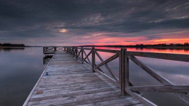 Polonya 'nın Mazury kentindeki sıcak ve renkli bir gökyüzü tarafından çerçevelenmiş, gün doğumunda sakin bir göle uzanan ahşap bir iskele..