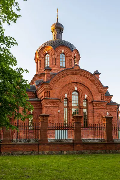Polonya 'nın Bialowieza şehrinde gün batımında dekoratif kuleleri ve kubbeleri olan kırmızı tuğlalı bir Ortodoks kilisesi çitin arkasında duruyor..