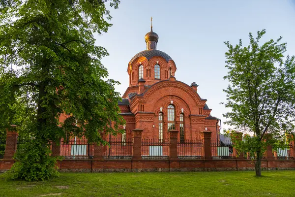 Polonya 'nın Bialowieza şehrinde gün batımında dekoratif kuleleri ve kubbeleri olan kırmızı tuğlalı bir Ortodoks kilisesi çitin arkasında duruyor..