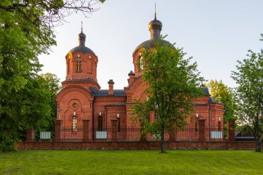 Polonya 'nın Bialowieza şehrinde gün batımında dekoratif kuleleri ve kubbeleri olan kırmızı tuğlalı bir Ortodoks kilisesi çitin arkasında duruyor..