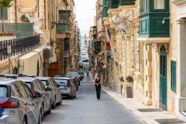 Valletta, Malta 21 Haziran 2021: Polis arabaları, eski binalar, balkonlar ve Valletta tarihi merkezinde yürüyen bir kadın ile dar eğimli cadde.