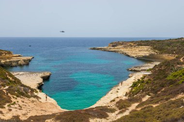 Forti Delimara, Malta 22 Haziran 2021: Rocky kıyı şeridi ve turkuaz körfezi insanların güneşlendiği ve helikopterle gökyüzünde uçtuğu yer.
