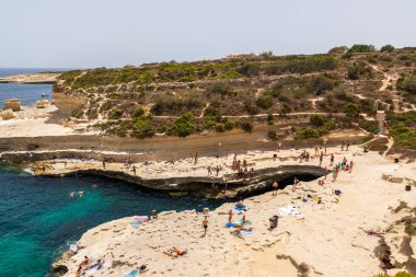 Aziz Peter Havuzu, Malta 22 Haziran 2021: Turistler açık turkuaz deniz kıyısında kayalık teraslarda yüzüyor ve güneşleniyor.