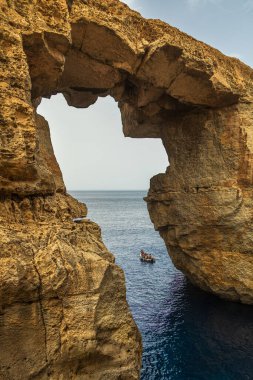 Gozo, Malta 23 Haziran 2021: Engebeli sahil boyunca uzanan doğal bir kaya kemerinin altındaki küçük bir bot geçidinde turistler.