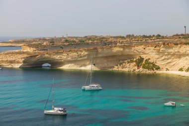 Taht Il-Maqjel, Malta 24 Haziran 2021: Arka planda kireçtaşı kemer ve engebeli kıyı şeridi bulunan turkuaz sulara demirlenmiş yatların yayın manzarası.