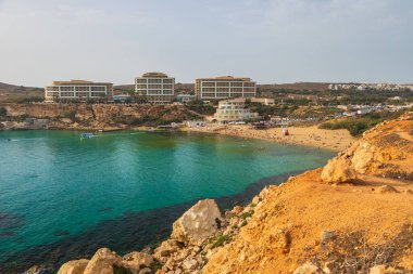 Manikata, Malta 24 Haziran 2021: Turkuaz sular ve çevre tepelerle Golden Bay sahilinde dinlenen turistler.