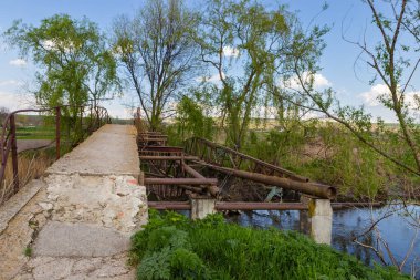Köydeki bir nehrin üzerinde yıkılmış eski bir köprü. Beton ve çelik köprü, yeşil ağaçlar, bahar zamanı, bulutlu mavi gökyüzü. Kırsal bölgede hasar gören köprü.