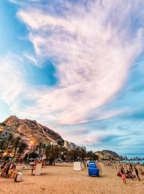 ALICANTE, İSPAN - 3 HAZİRAN 2023 Playa del Postiguet plajında akşam vakti, arka planda Santa Barbara kalesi. Palmiye ağaçları ve güzel mavi ve pembe bulutlu gökyüzü, dikey çekim.