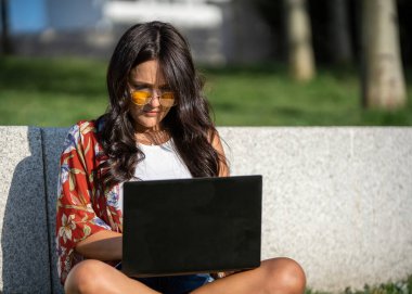 Parkta çimlerin üzerinde otururken güneş gözlüklü bir kadının dizüstü bilgisayar kullanışının yakın görüntüsü. Teknoloji konsepti.