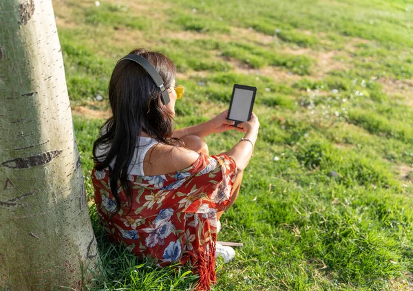 Woman with headphones reading an e-book with a blank screen while sitting outdoors on grass. Technology concept.