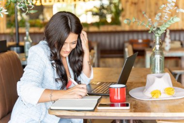Kahve dükkanında dizüstü bilgisayar kullanan, gülümseyen ve not defterine yazan bir kadın. Teknoloji, yaratıcılık ve iş konsepti.