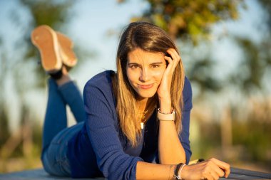 Gün batımında bir bankta uzanmış kameraya bakıp gülümseyen bir kadın..