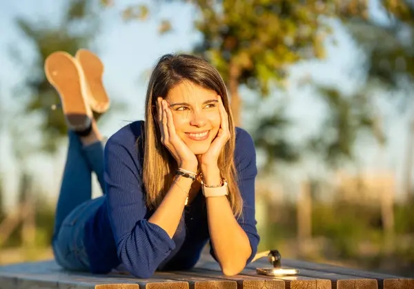Happy and relaxed woman smiling while lying on a bench outdoors in a park during the sunset.