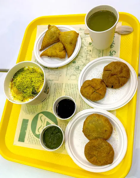 Masoori, Hintli, 30 Nisan 2024: Sarı tepside servis edilen geleneksel Hint sokak yemeklerinin canlı bir çeşidi. Görüntüde kachori, samosa, bir kase sev bile puri ve bir fincan yeşil baharatlı içecek yer alıyor.