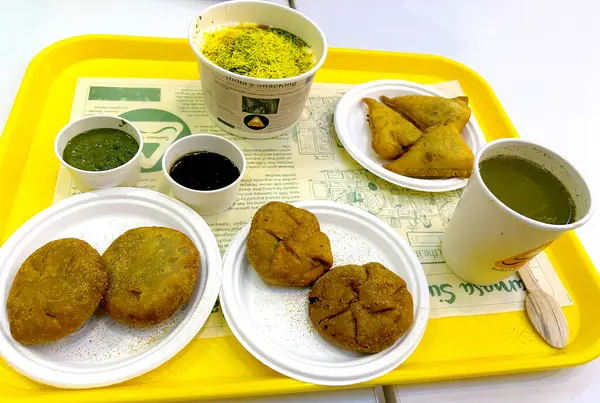 Masoori, Hintli, 30 Nisan 2024: Sarı tepside servis edilen geleneksel Hint sokak yemeklerinin canlı bir çeşidi. Görüntüde kachori, samosa, bir kase sev bile puri ve bir fincan yeşil baharatlı içecek yer alıyor.