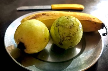 İki taze guava ve arkaplanda bir bıçakla geleneksel çelik bir tabağa yerleştirilmiş olgun bir muzun yakın plan görüntüsü.