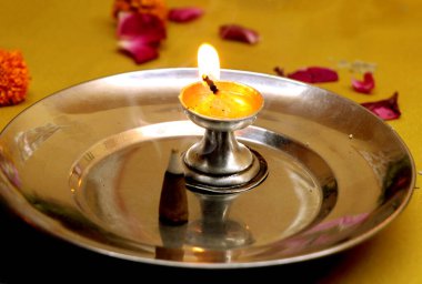 Parlayan bir yağ lambası gümüş bir tabağa konmuş, yanında da kadife çiçekleri ve gül yapraklarıyla çevrili, Hindu dini ayinleri ve kutlamalarında saflığı, bağlılığı ve ışığı simgeleyen bir tütsü.