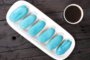 Beyaz dikdörtgen bir plakaya dizilmiş altı canlı mavi Clair 'in yakın çekimi. Clair 'lerin parlak bir kapanışı var ve mükemmel bir şekilde hizalanmışlar. Tabağın yanındaki ahşap masaya bir fincan kahve konur, tatlı ve lezzetli kombinasyonu tamamlar.. 