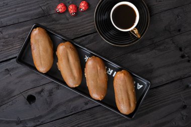 Siyah dikdörtgen bir tabakta dört karamel kaplı ekin ve yanında da altın kaplamalı siyah bir fincan kahve. Sahne, üç dekoratif kırmızı uğur böceğiyle birlikte koyu ahşap bir masada geçiyor..