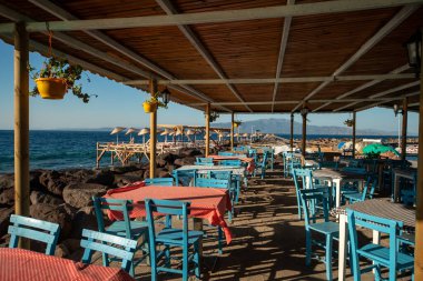 Geleneksel Akdeniz Sahil Restoranı Mavi Tahta Sandalyeler ve Deniz Kenarı Terasında Kırmızı Kareli Masa örtüleri Yaz boyunca Rocky Coast manzaralı.