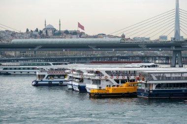 03.13.2025, İstanbul, Türkiye. Modern bir köprünün yakınına yanaşmış tekneleri olan sahile bakan bulutlu bir gökyüzünün altında tarihi bir cami, gelenek ve kentsel yaşamı harmanlayan bir şehir manzarası sergileniyor..