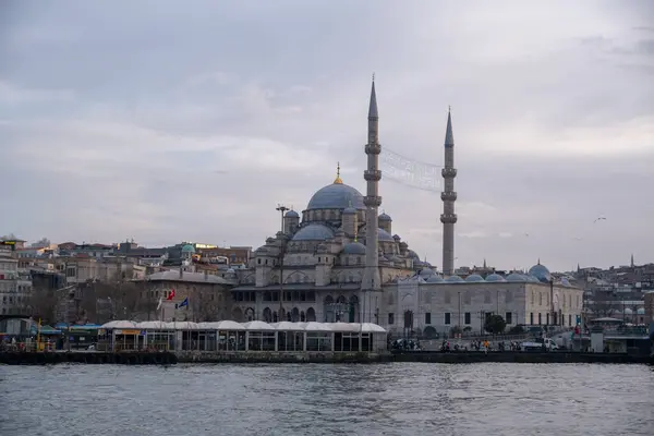 Çarpıcı kubbeleri ve minareleri olan görkemli bir cami, dramatik bulutlu bir gökyüzü altında tarihi bir şehir manzarası tarafından çerçevelenmiş sakin bir rıhtıma bakıyor..