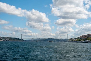 İstanbul, Türkiye 'deki Boğaz Köprüsü' nün altından geçen büyük gemilerle İstanbul 'da balıkçı tekneleri, manzaralı bulutlar, tepeler ve arka planda bir Türk bayrağıyla aydınlık bir günde