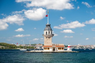 İstanbul 'daki tarihi Bakire Kulesi' nde güneşli bir günde rıhtıma yanaşan gezici tekneler, Boğaziçi suları, mavi gökyüzü, şehir silüeti ve ikonik Türk mimarisi yer alıyor..