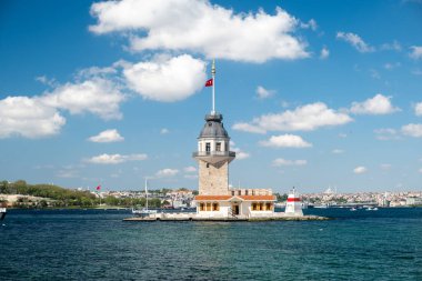 İstanbul 'daki tarihi Bakire Kulesi' nde güneşli bir günde rıhtıma yanaşan gezici tekneler, Boğaziçi suları, mavi gökyüzü, şehir silüeti ve ikonik Türk mimarisi yer alıyor..