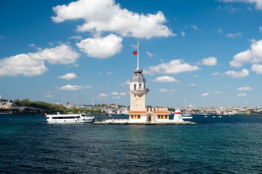 İstanbul 'daki tarihi Bakire Kulesi' nde güneşli bir günde rıhtıma yanaşan gezici tekneler, Boğaziçi suları, mavi gökyüzü, şehir silüeti ve ikonik Türk mimarisi yer alıyor..