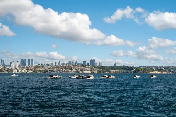 İstanbul, Türkiye 'deki Boğaz Köprüsü' nün altından geçen büyük gemilerle İstanbul 'da balıkçı tekneleri, manzaralı bulutlar, tepeler ve arka planda bir Türk bayrağıyla aydınlık bir günde