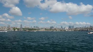 İstanbul, Türkiye 'deki Boğaz Köprüsü' nün altından geçen büyük gemilerle İstanbul 'da balıkçı tekneleri, manzaralı bulutlar, tepeler ve arka planda bir Türk bayrağıyla aydınlık bir günde